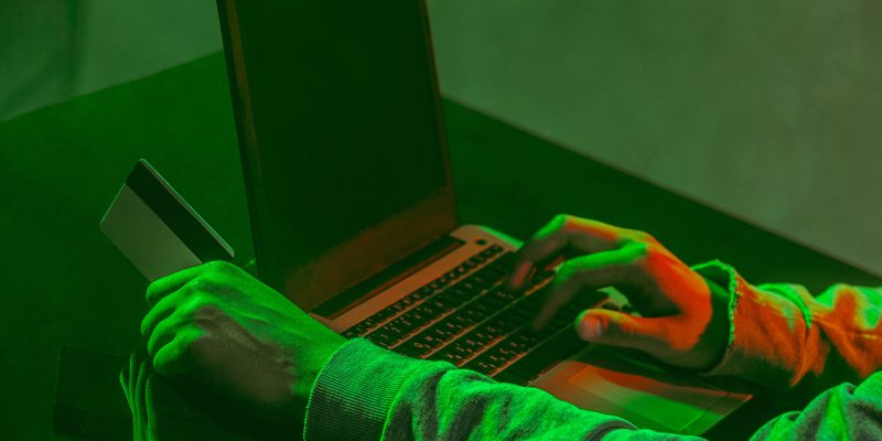Hooded computer hacker stealing information with laptop on colored studio background